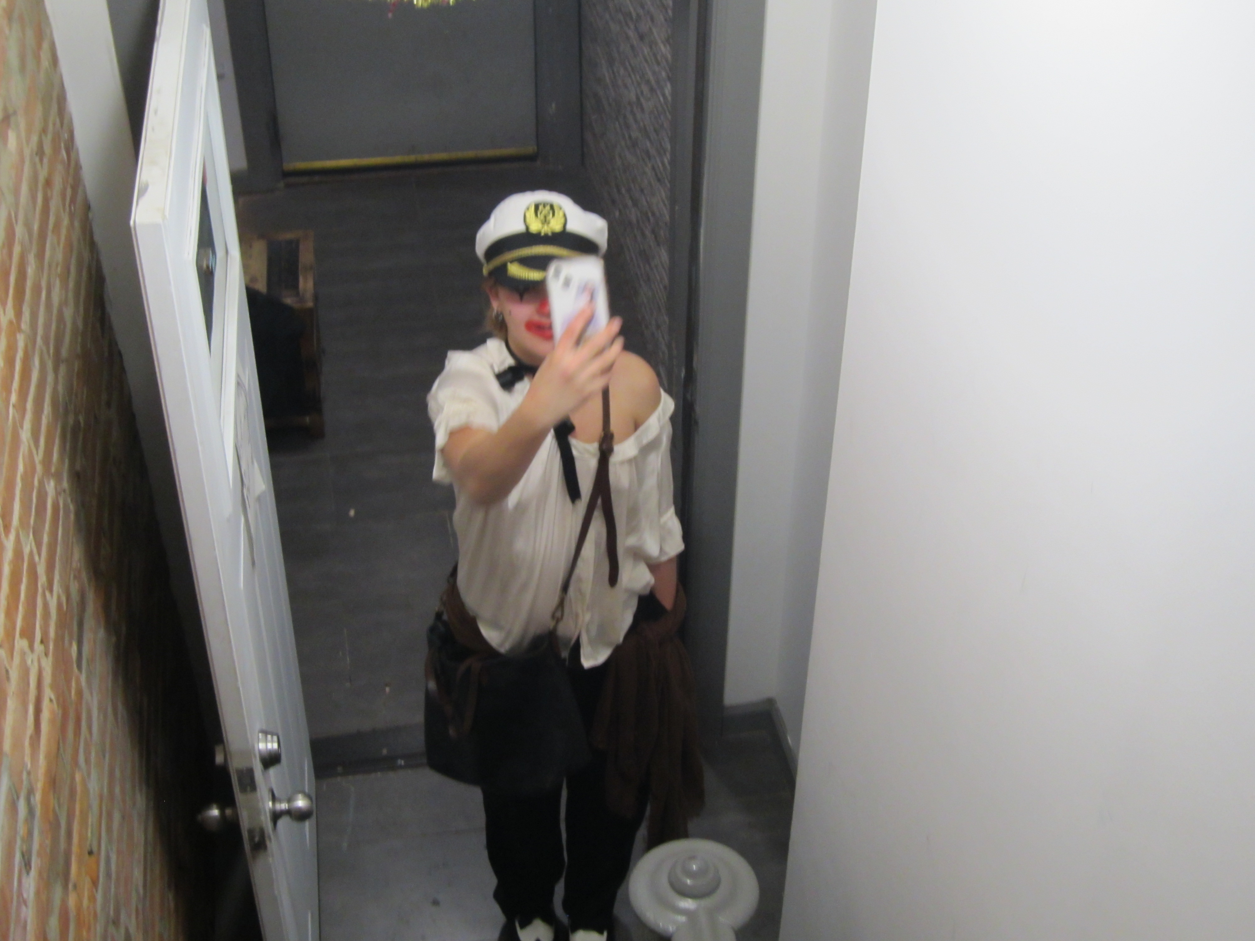 clown wearing a sailors hat and white silky blouse takes a selfie in front of a white doorway